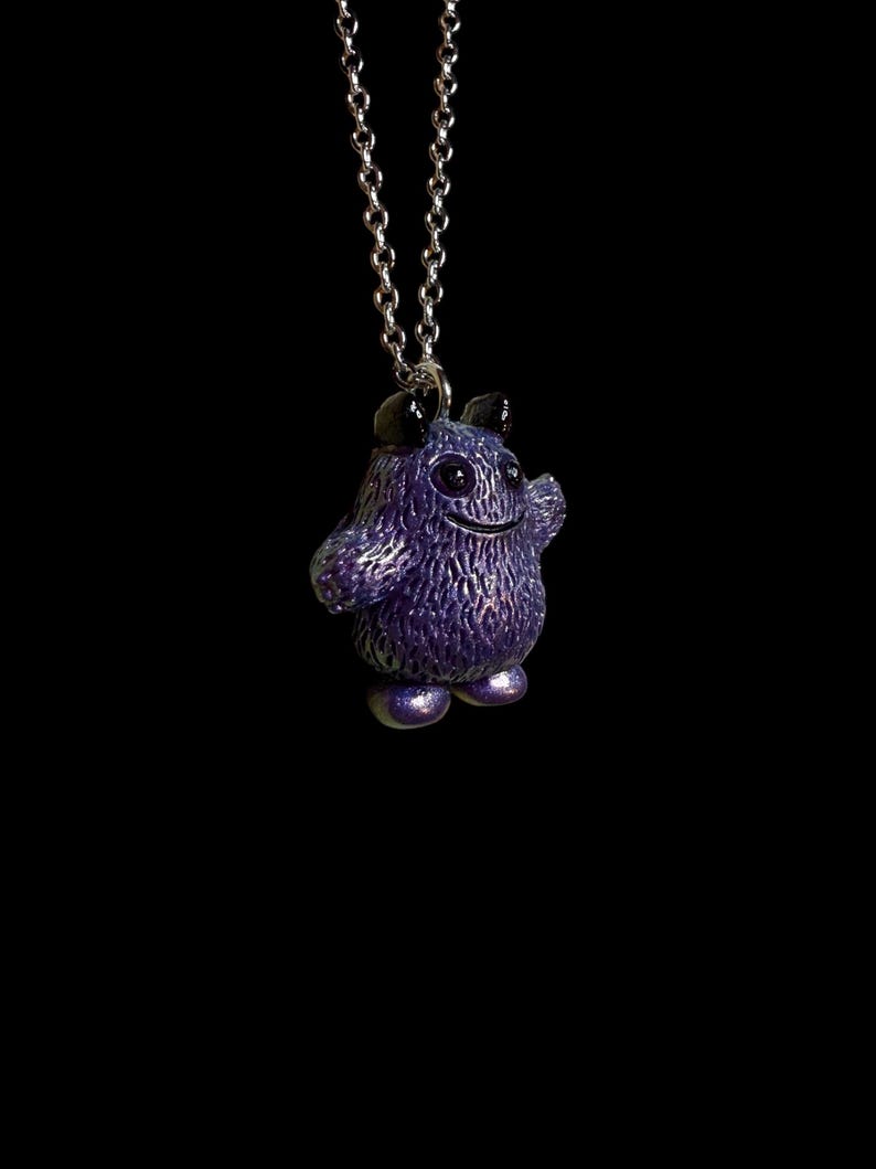 May include: A purple monster pendant on a silver chain. The pendant has a textured surface, small horns, and a smiling face. The monster is standing with its arm raised. The background is black.