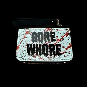 May include: A white zippered wallet with a black strap. The wallet has red blood splatter and the words "GORE WHORE" printed in black.