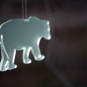 May include: A translucent, light green bear-shaped ornament suspended by a thin string. The bear is depicted in a walking pose, with visible details of its paws and body. The background is a blurred dark color.