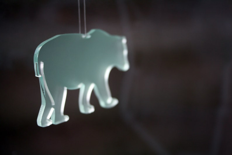 May include: A translucent, light green bear-shaped ornament suspended by a thin string. The bear is depicted in a walking pose, with visible details of its paws and body. The background is a blurred dark color.