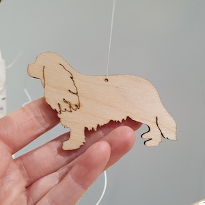 May include: A wooden ornament shaped like a Cavalier King Charles Spaniel dog. The ornament is light brown and has a string attached for hanging.
