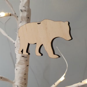 May include: A wooden bear ornament, cut out in the shape of a bear, with a natural wood finish. The ornament is hanging from a white string.