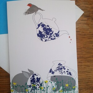 May include: A greeting card with a blue and white teapot with a red robin perched on top, a gray rabbit wearing a blue and white teacup, and a second gray rabbit wearing a blue and white teacup in a field of green grass and yellow flowers.