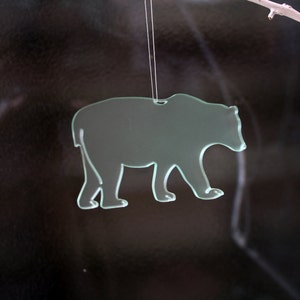 May include: A translucent glass bear ornament, suspended by a thin string. The bear is depicted in a walking pose, with a cutout design. The background is dark, with a white branch in the upper right corner.