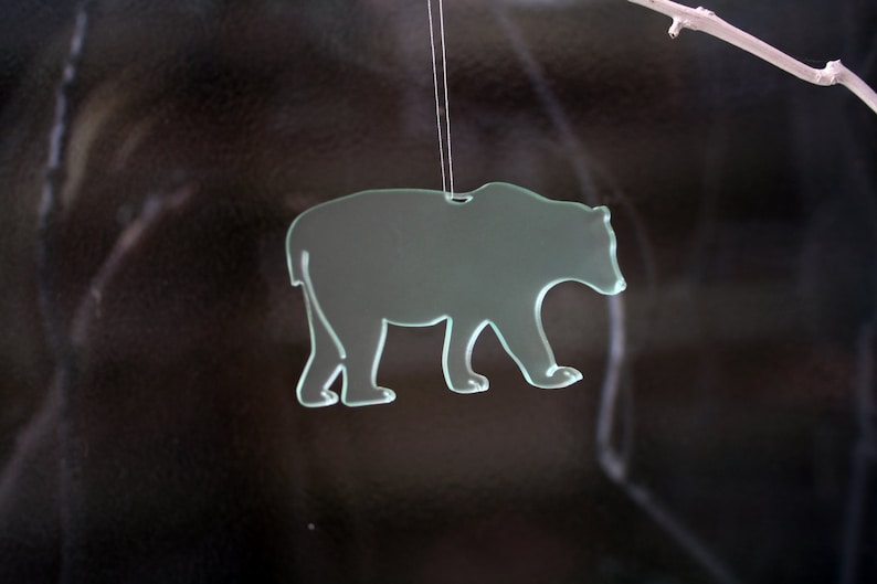 May include: A translucent glass bear ornament, suspended by a thin string. The bear is depicted in a walking pose, with a cutout design. The background is dark, with a white branch in the upper right corner.