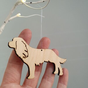 May include: A wooden dog ornament, cut from light-colored wood, hangs from a thin white string. The dog's silhouette is detailed with cut-out features and a dark edge. The background is a soft gray.