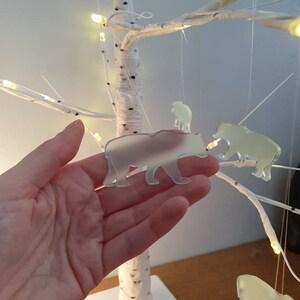 May include: A decorative birch tree with hanging bear ornaments. The bears are translucent and suspended by thin strings. The tree has white branches and a square base. Small lights are wrapped around the branches.