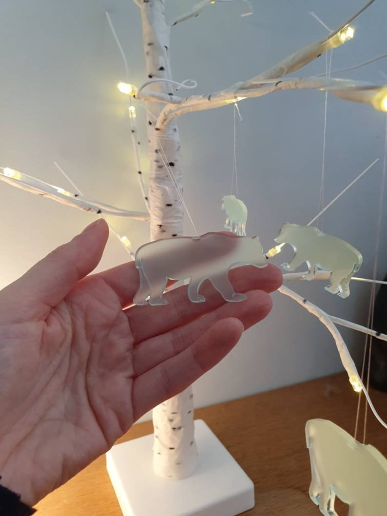 May include: A decorative birch tree with hanging bear ornaments. The bears are translucent and suspended by thin strings. The tree has white branches and a square base. Small lights are wrapped around the branches.
