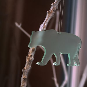 May include: A translucent green bear-shaped ornament hanging from a thin string. The bear is suspended from a small, textured branch. The background is blurred, with hints of brown and white.