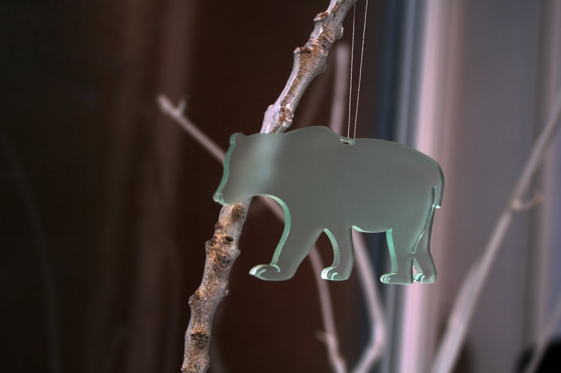 May include: A translucent green bear-shaped ornament hanging from a thin string. The bear is suspended from a small, textured branch. The background is blurred, with hints of brown and white.