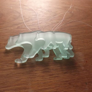 May include: Three frosted glass bear ornaments with attached strings. The bears are in a walking pose, with the details of their paws and bodies visible. The ornaments are a pale green color and are displayed on a wooden surface.