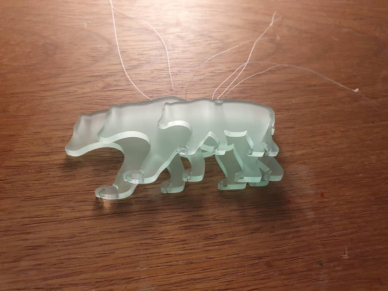 May include: Three frosted glass bear ornaments with attached strings. The bears are in a walking pose, with the details of their paws and bodies visible. The ornaments are a pale green color and are displayed on a wooden surface.