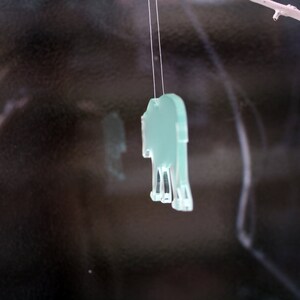 May include: A light green, transparent elephant-shaped ornament hanging from a thin string. The ornament is suspended in front of a dark, reflective background. A small branch is visible in the upper right corner.
