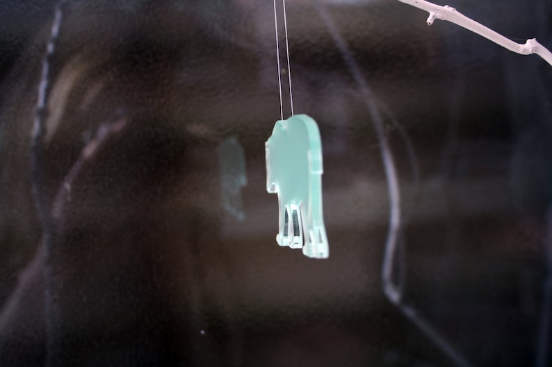 May include: A light green, transparent elephant-shaped ornament hanging from a thin string. The ornament is suspended in front of a dark, reflective background. A small branch is visible in the upper right corner.