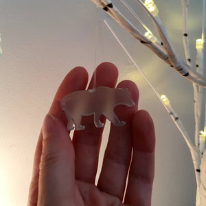 May include: A translucent bear-shaped ornament suspended by a thin string. The bear is held in front of a white, illuminated decorative tree. The ornament is a light, neutral color, and the tree has warm white lights.
