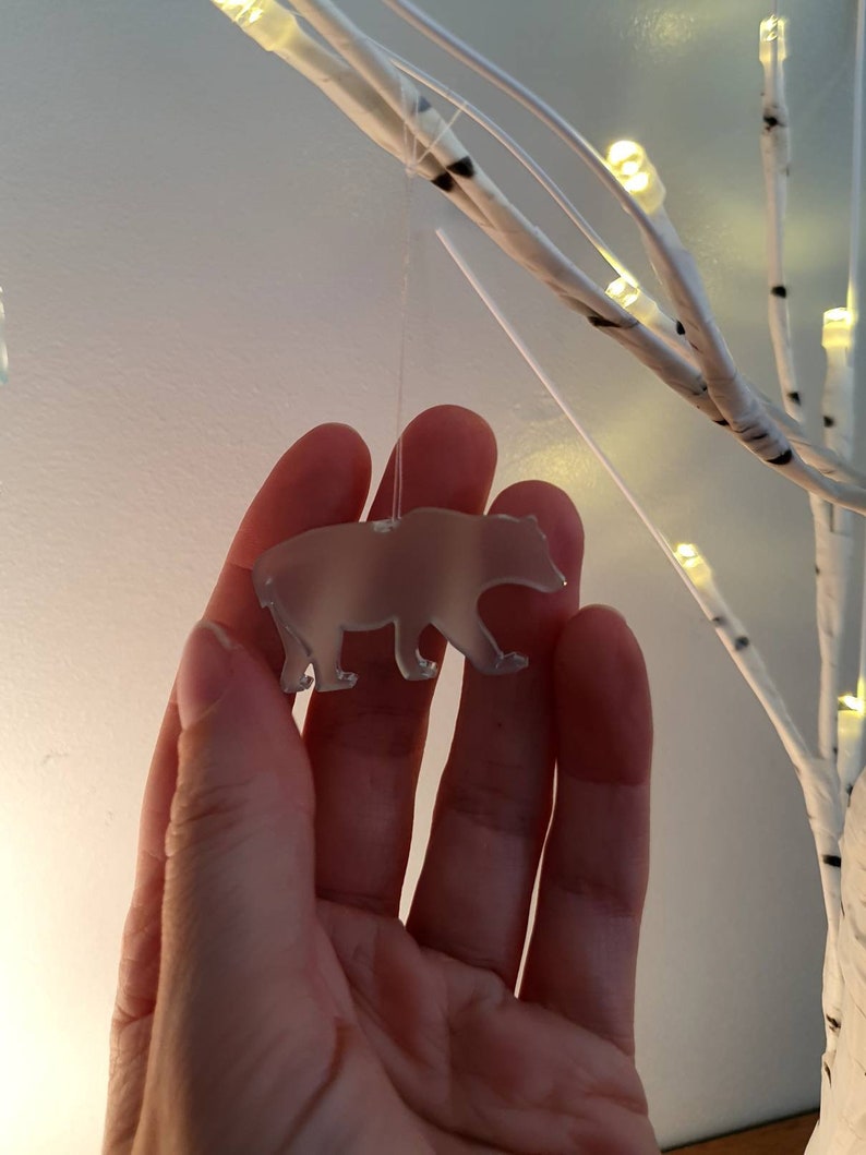 May include: A translucent bear-shaped ornament suspended by a thin string. The bear is held in front of a white, illuminated decorative tree. The ornament is a light, neutral color, and the tree has warm white lights.