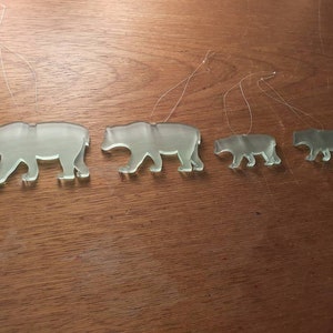 May include: Four clear glass bear ornaments of varying sizes are displayed. Each bear is in a walking pose and has a thin string attached for hanging. The ornaments are arranged from largest to smallest, showcasing a family theme.