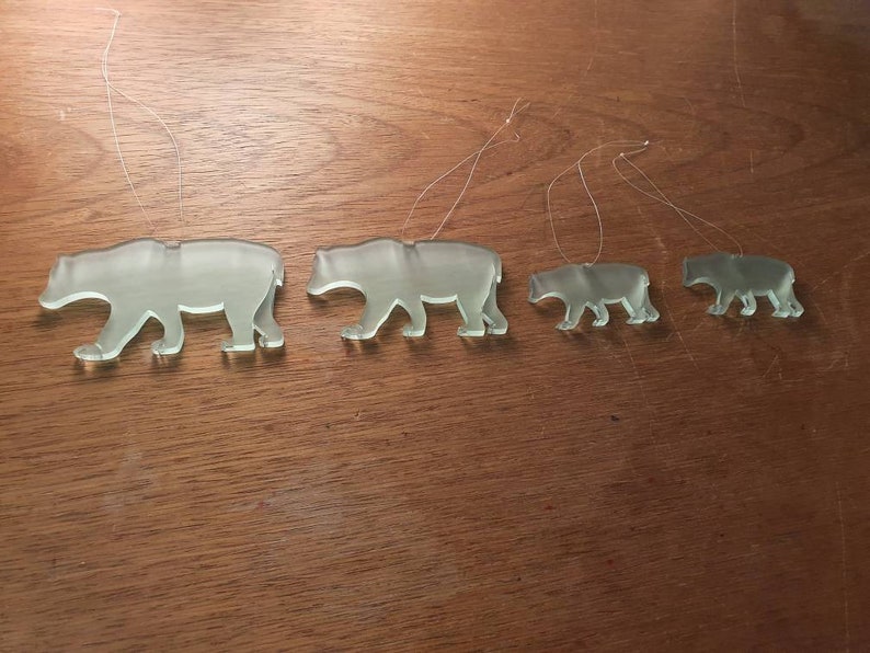 May include: Four clear glass bear ornaments of varying sizes are displayed. Each bear is in a walking pose and has a thin string attached for hanging. The ornaments are arranged from largest to smallest, showcasing a family theme.