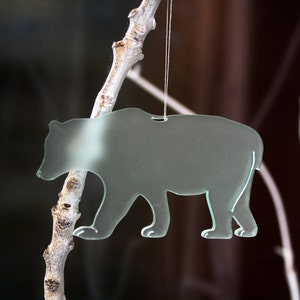 May include: A clear glass bear ornament hanging from a thin string. The bear is walking, with its shape outlined in white. The background is blurred, with a dark brown and white color scheme.