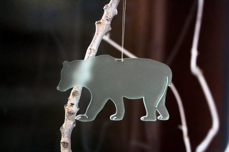 May include: A clear glass bear ornament hanging from a thin string. The bear is walking, with its shape outlined in white. The background is blurred, with a dark brown and white color scheme.