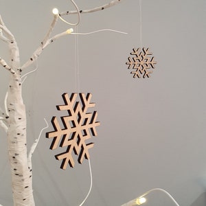 May include: Two wooden snowflake ornaments hanging from a white birch branch with white lights. The snowflakes are different sizes and are made of natural wood.