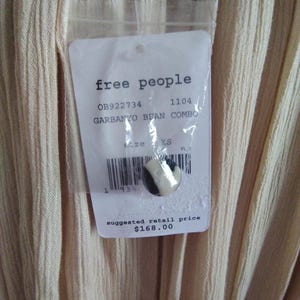 May include: Close-up of a garment with a tag from "free people". The tag includes the text "GARBANZO BEAN COMBO" and "XS" size. The garment is a light beige color with vertical ribbed texture.