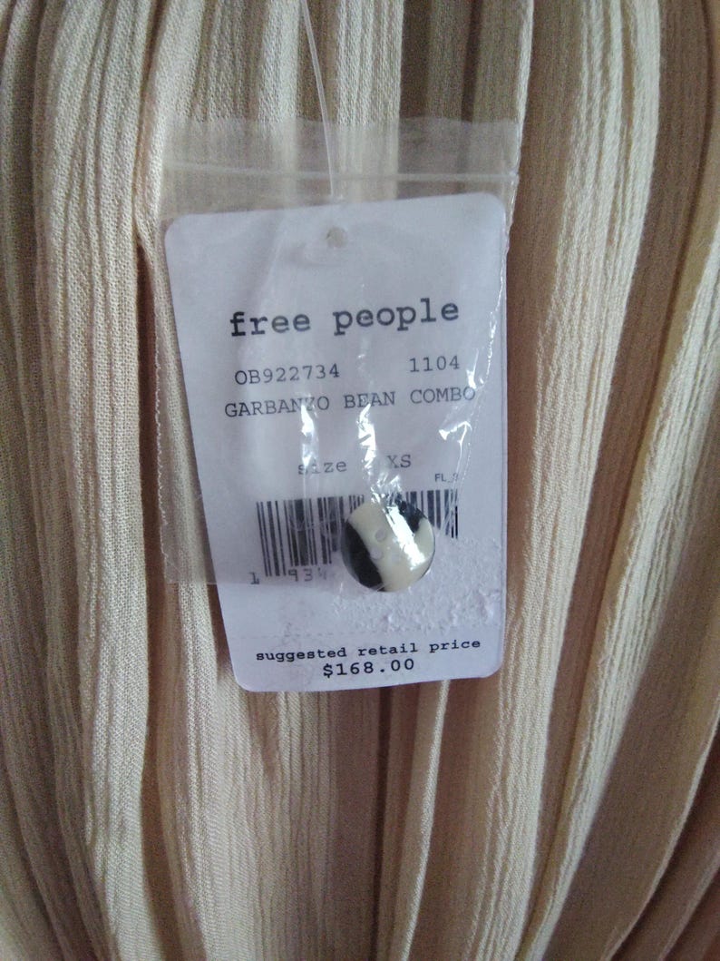 May include: Close-up of a garment with a tag from "free people". The tag includes the text "GARBANZO BEAN COMBO" and "XS" size. The garment is a light beige color with vertical ribbed texture.