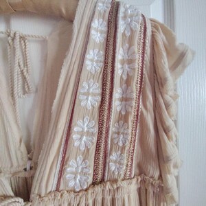 May include: Cream-colored dress with white floral embroidery and red and gold accents. The dress features a V-neckline and ruffled sleeves. The fabric appears lightweight and flowing, suitable for warm weather. The dress is hanging on a wooden hanger.