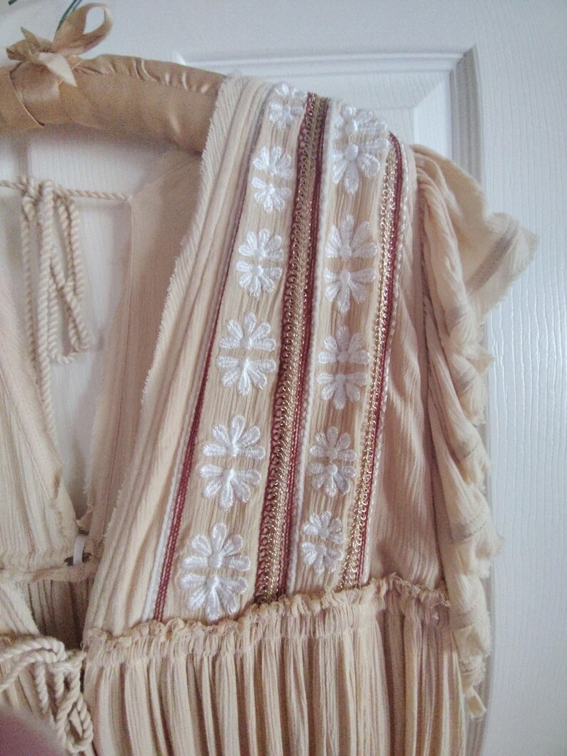 May include: Cream-colored dress with white floral embroidery and red and gold accents. The dress features a V-neckline and ruffled sleeves. The fabric appears lightweight and flowing, suitable for warm weather. The dress is hanging on a wooden hanger.