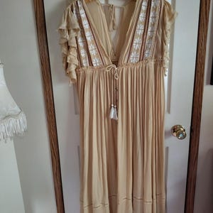May include: A beige, flowing dress with a deep V-neck and short, ruffled sleeves. The bodice features white and red embroidered floral details. The dress is hanging on a wooden hanger and has a tassel tie at the waist. The dress is approximately knee-length.
