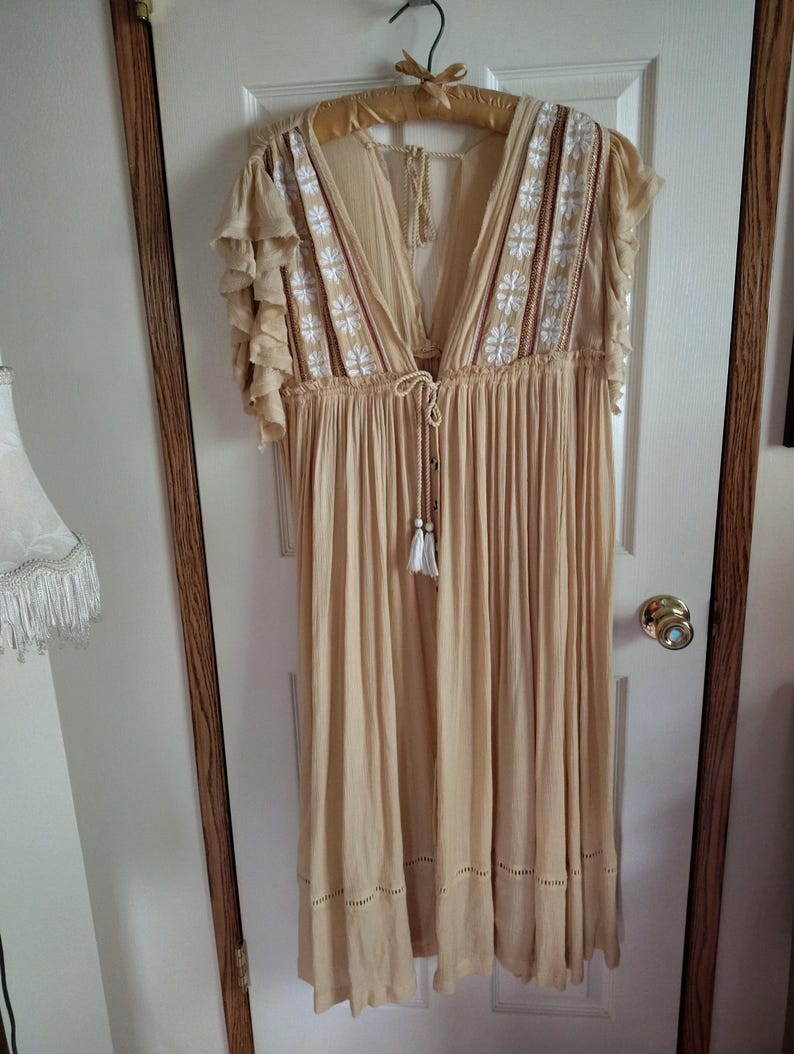 May include: A beige, flowing dress with a deep V-neck and short, ruffled sleeves. The bodice features white and red embroidered floral details. The dress is hanging on a wooden hanger and has a tassel tie at the waist. The dress is approximately knee-length.