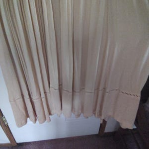 May include: A flowing, light beige dress with vertical pleats. The hemline features a decorative, openwork border. The fabric appears lightweight and suitable for warm weather. The dress hangs from the top of the frame.