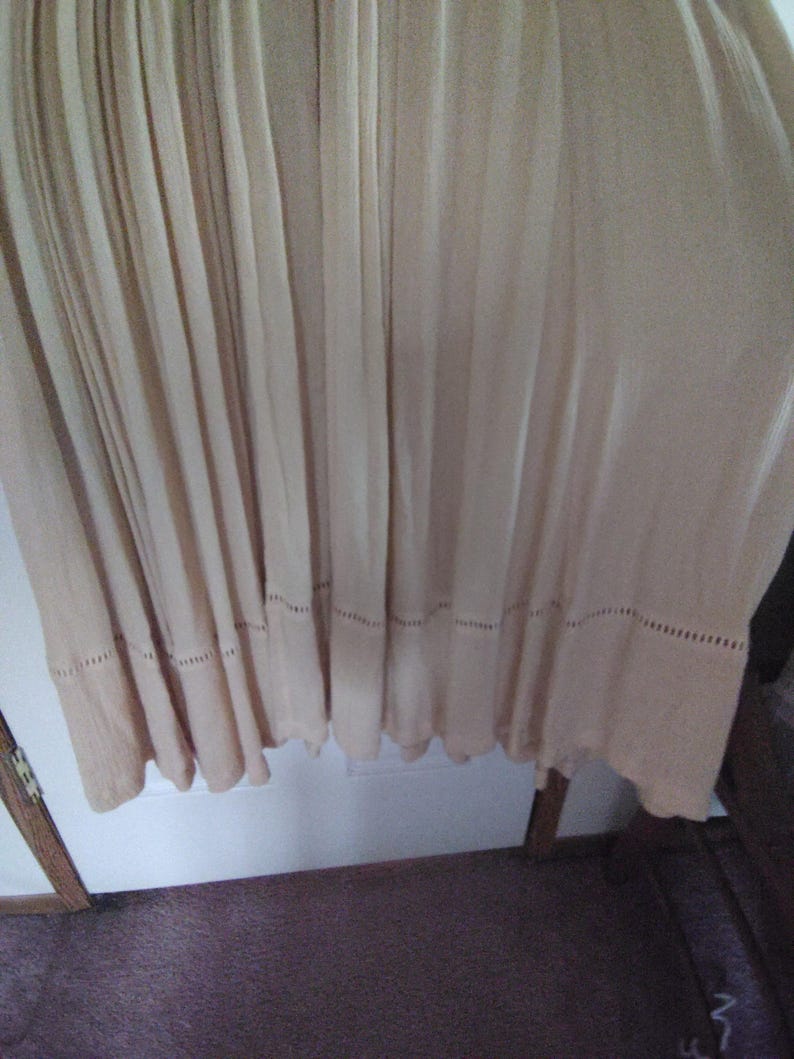 May include: A flowing, light beige dress with vertical pleats. The hemline features a decorative, openwork border. The fabric appears lightweight and suitable for warm weather. The dress hangs from the top of the frame.