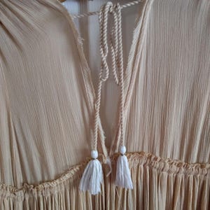 May include: A light peach-colored dress with a textured fabric and a V-neckline. The dress features a drawstring with white tassels and a ruffled detail at the waist. The dress is hanging on a wooden hanger.