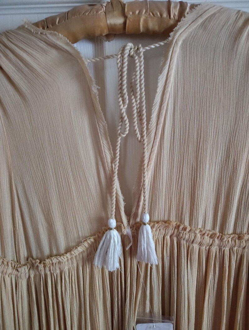 May include: A light peach-colored dress with a textured fabric and a V-neckline. The dress features a drawstring with white tassels and a ruffled detail at the waist. The dress is hanging on a wooden hanger.