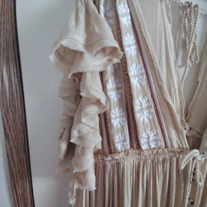May include: A beige dress with ruffled sleeves and a floral embroidered bodice. The dress is hanging on a wooden hanger. The dress has a flowing skirt and a drawstring neckline.