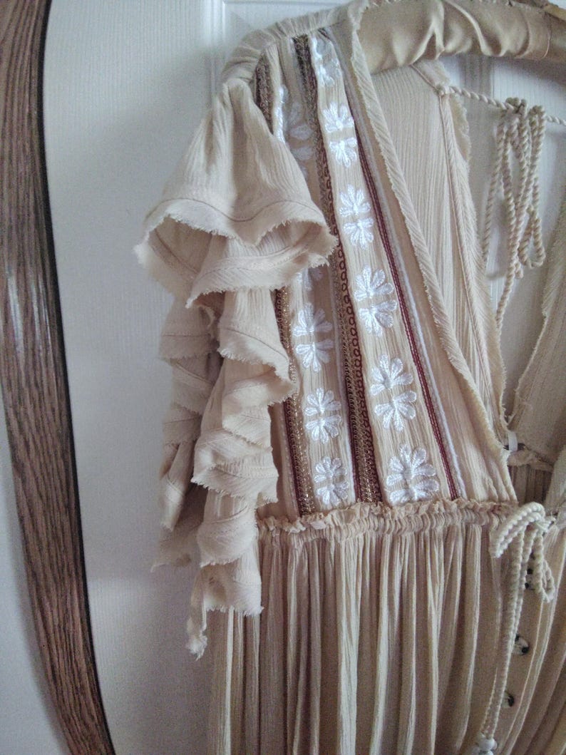 May include: A beige dress with ruffled sleeves and a floral embroidered bodice. The dress is hanging on a wooden hanger. The dress has a flowing skirt and a drawstring neckline.