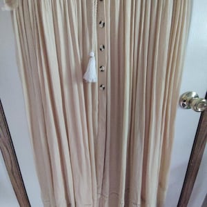 May include: Off-the-shoulder, cream-colored dress with a button-down front and a tassel detail. The dress features a drawstring neckline and a scalloped hemline. The fabric has a pleated texture, creating a flowing silhouette.