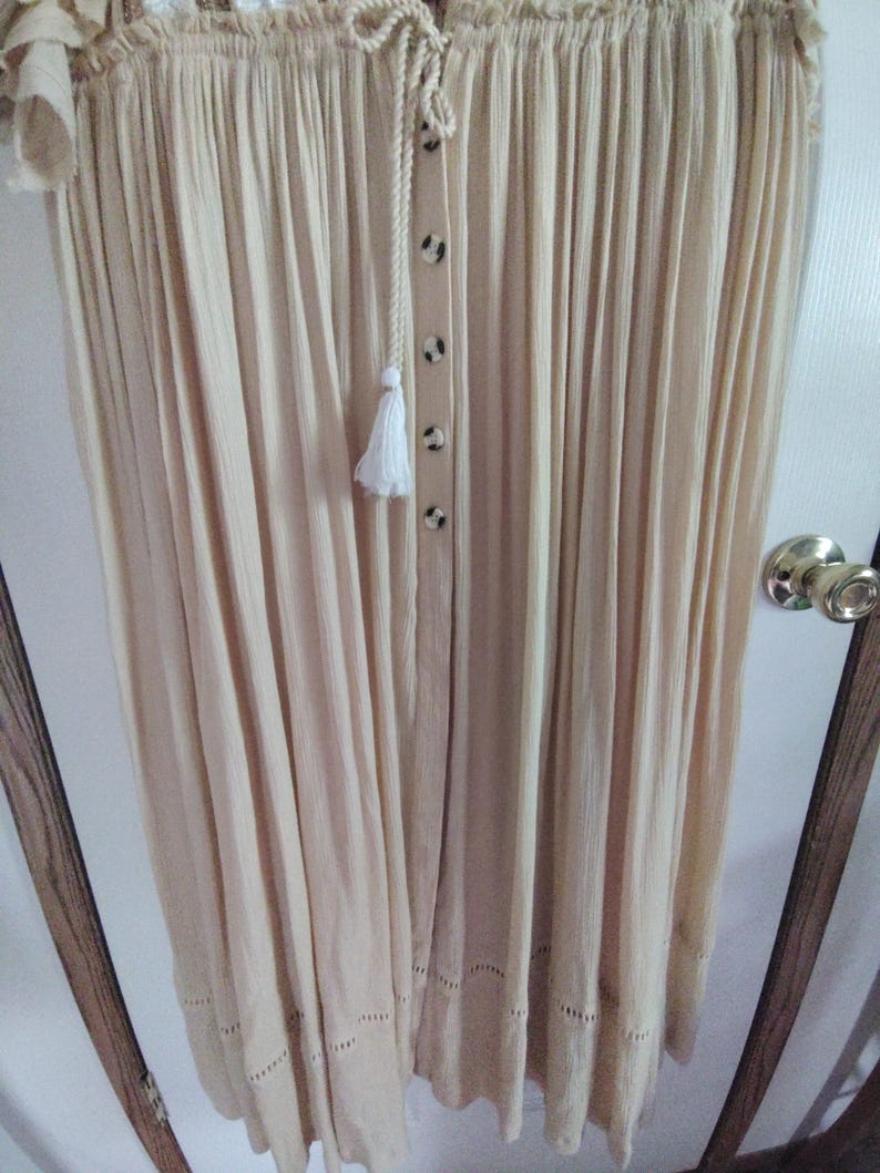May include: Off-the-shoulder, cream-colored dress with a button-down front and a tassel detail. The dress features a drawstring neckline and a scalloped hemline. The fabric has a pleated texture, creating a flowing silhouette.
