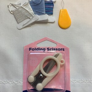 Puede incluir: Un paquete de tijeras plegables blancas sobre un fondo rosa y azul. El paquete dice "Folding Scissors" y "Hemline".