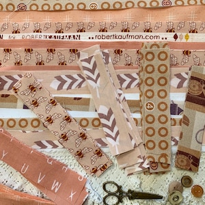 Puede incluir: Una colección de tiras de tela con varios patrones y colores, incluyendo abejas, plumas y círculos. Las telas son en tonos beige, marrón y crema. También se incluyen tijeras, botones y tela con el texto "robertkaufman.com".