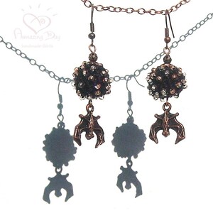 May include: A pair of earrings with black and silver bead clusters and dangling bat charms. The earrings are attached to a silver chain necklace.