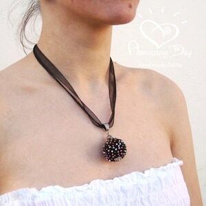 May include: A black ribbon necklace with a silver pendant featuring a round, beaded ornament in shades of black, purple, and silver. The pendant is attached to the ribbon with a silver loop. The necklace is worn by a person with a white strapless top.