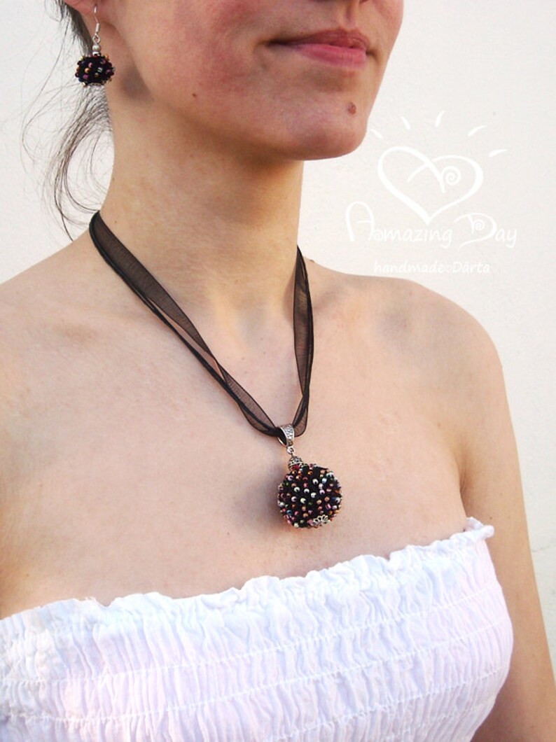 May include: A black ribbon necklace with a silver pendant featuring a round, beaded ornament in shades of black, purple, and silver. The pendant is attached to the ribbon with a silver loop. The necklace is worn by a person with a white strapless top.