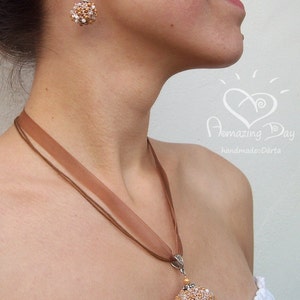 May include: A brown ribbon necklace with a beaded pendant in the shape of a sphere. The pendant is made of gold and white beads. The necklace is worn by a person with a pair of earrings that match the pendant.