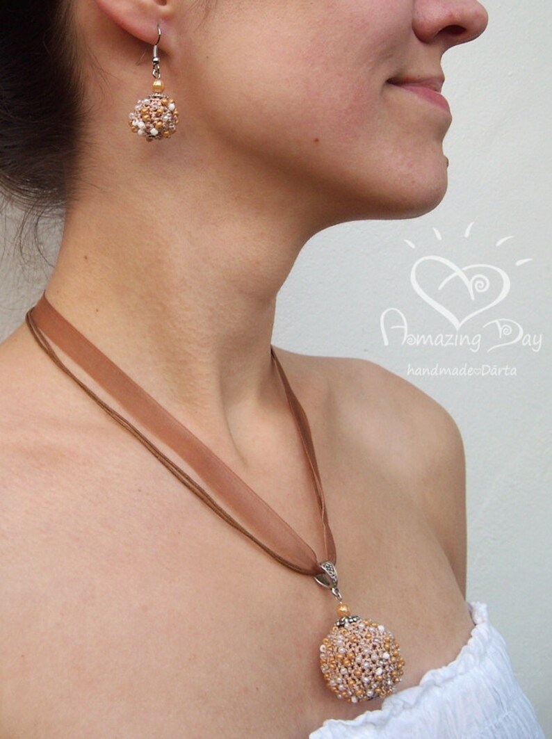 May include: A brown ribbon necklace with a beaded pendant in the shape of a sphere. The pendant is made of gold and white beads. The necklace is worn by a person with a pair of earrings that match the pendant.