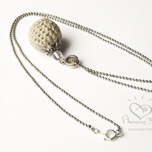 May include: A silver chain necklace with a crocheted beige ball pendant. The pendant has a silver embellishment and a clear bead.