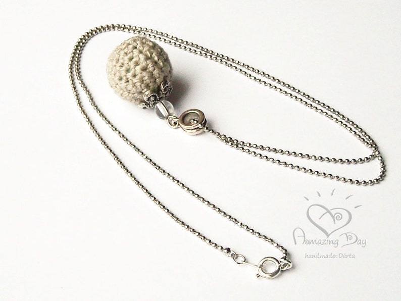 May include: A silver chain necklace with a crocheted beige ball pendant. The pendant has a silver embellishment and a clear bead.