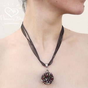 May include: A black ribbon necklace with a round, beaded pendant. The pendant is made of black, red, and gold beads. The necklace is worn by a person with a white shirt.
