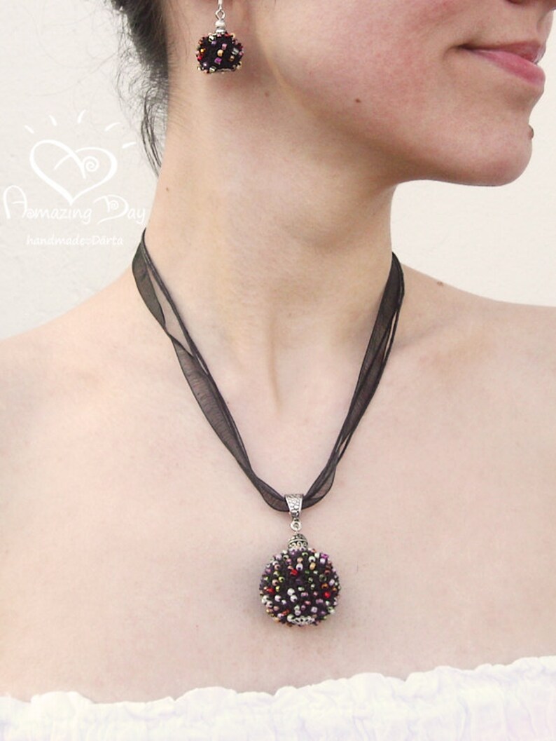 May include: A black ribbon necklace with a round, beaded pendant. The pendant is made of black, red, and gold beads. The necklace is worn by a person with a white shirt.